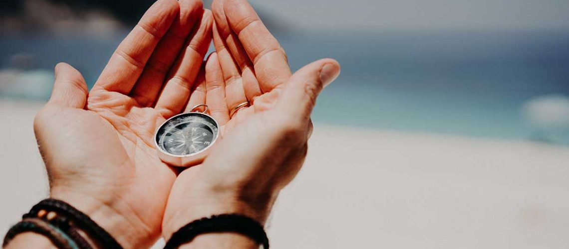 mans-hand-holding-compass-against-the-backdrop-of-beach-and-sea-the-concept-of-travel_1200x628